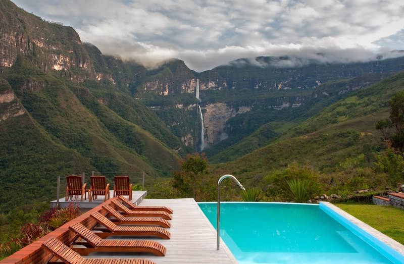 It bears repeating—this view is simply spectacular! Best Hikes In Peru Image: A hotel swimming pool is surrounded by wooden lounge chairs, and offers an incredible view of the waterfall at the other end of this mountainous landscape.