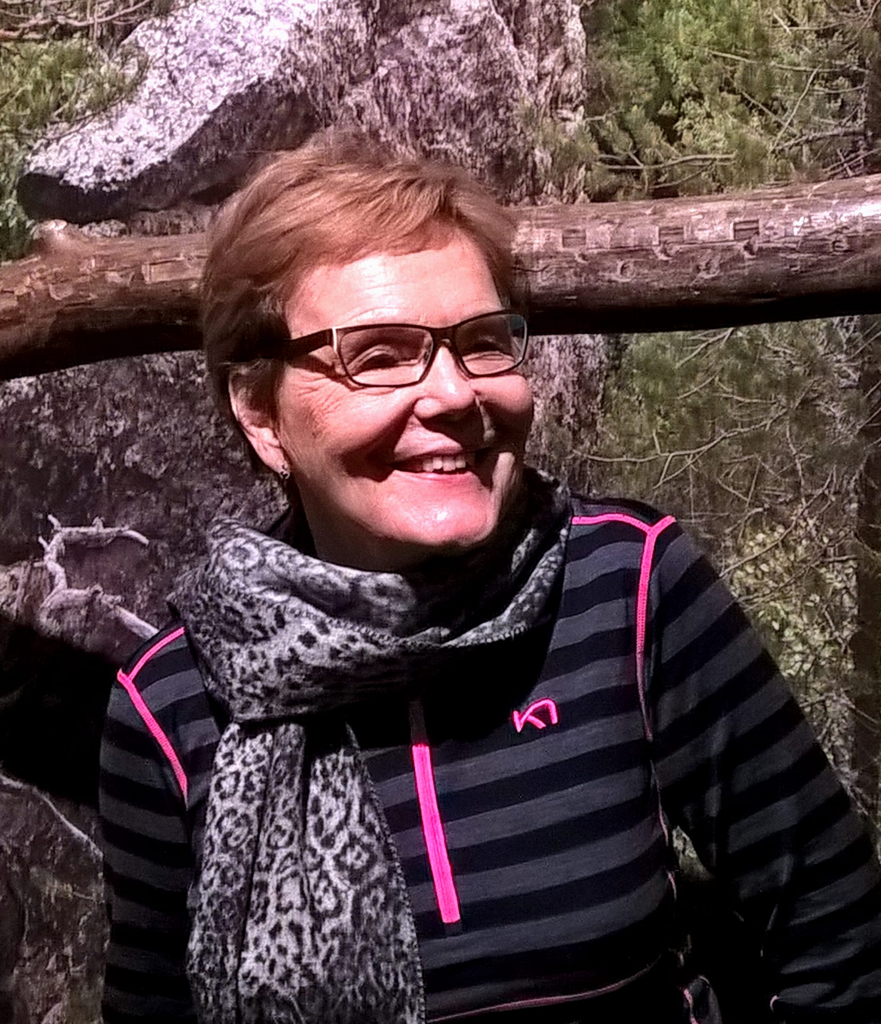 A photograph of author, Irmeli Westermarck. Seen wearing glasses, a leopard print scarf, and a grey and black striped shirt with hot pink accents.
