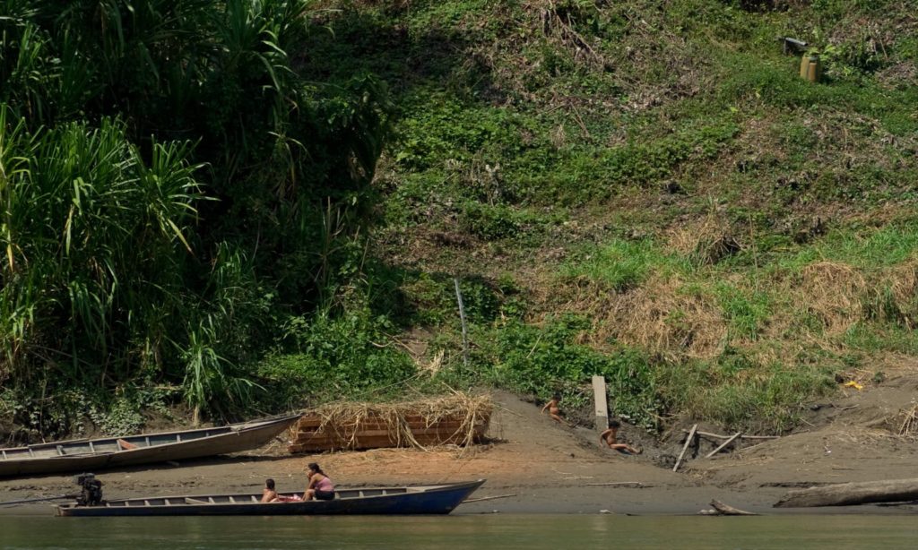 Rainforests & Natural Wonders: Manu National Park. Seen are people who may be members of an indigenous tribe sitting on the edge of a shore, and in a long narrow boat.