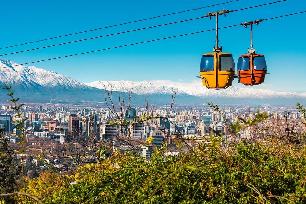 Destinations Worth Dreaming Image: An enclosed aerial tram has two pods: one yellow, one orange. They are sailing above the city of Chile; a snow-capped mountain range sits in the background, the city skyline is below, and greenery graces the foreground.