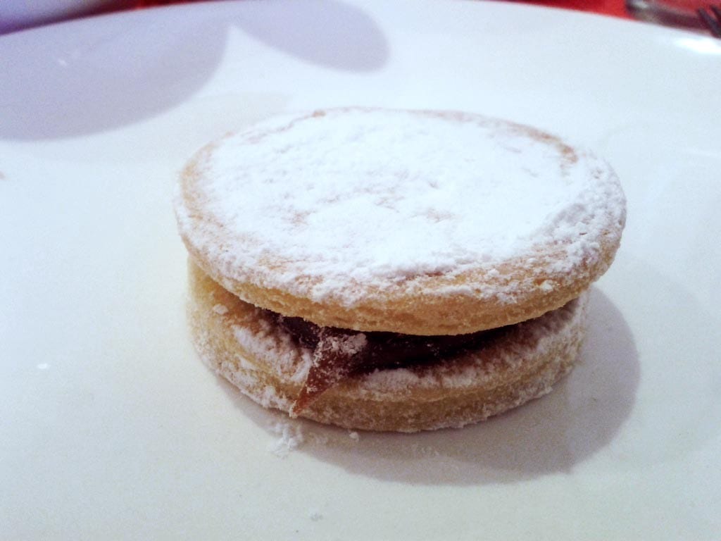 Visit Peru Image: A single alfajores sits on a white plate, and has been dusted with a generous amount of powdered (or confectioner's) sugar.