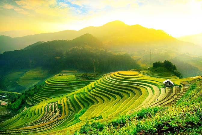Destinations Worth Dreaming Image: A green hill with sparse cottages in the mountains descends into a series of well-groomed levels sloping down to the valley below. 
