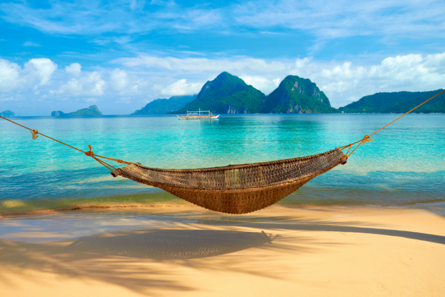Spend your holiday however you want. Want to lounge on the beach? We know the best beachside accommodations. Booking A Vacation Online Image: A hammock is suspended on a tropical beach over golden sand. Clear turquoise waters, a blue and white sky, and green mountainous islands are seen in the distance.