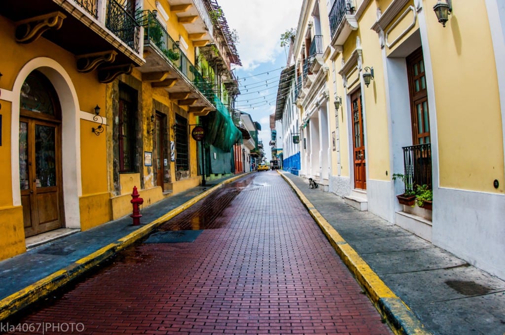 Architecture In Panama Image: A narrow city street is lined with buildings on both the left and the right. We can see shades of mustard yellow, pale lemon, turquoise, and marine blue gracing their faces. Many have wrought iron balconies.