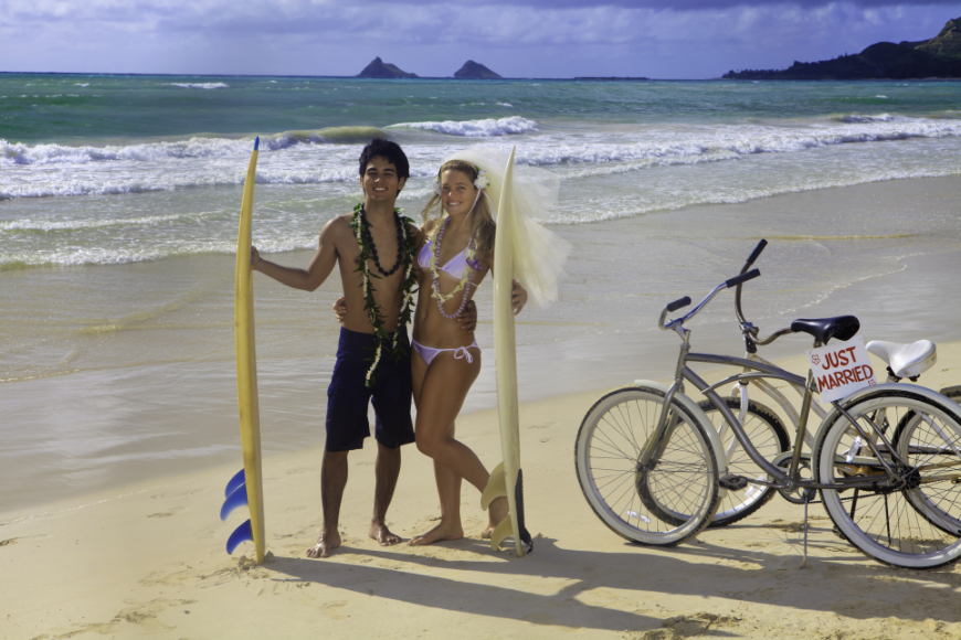 Your destination wedding can be as unique as you want it to be — just be sure to keep it green.  Eco-Friendly Destination Wedding Image: Newlyweds pose in swimsuits holding yellow surfboards; bikes are parked off to the side.
