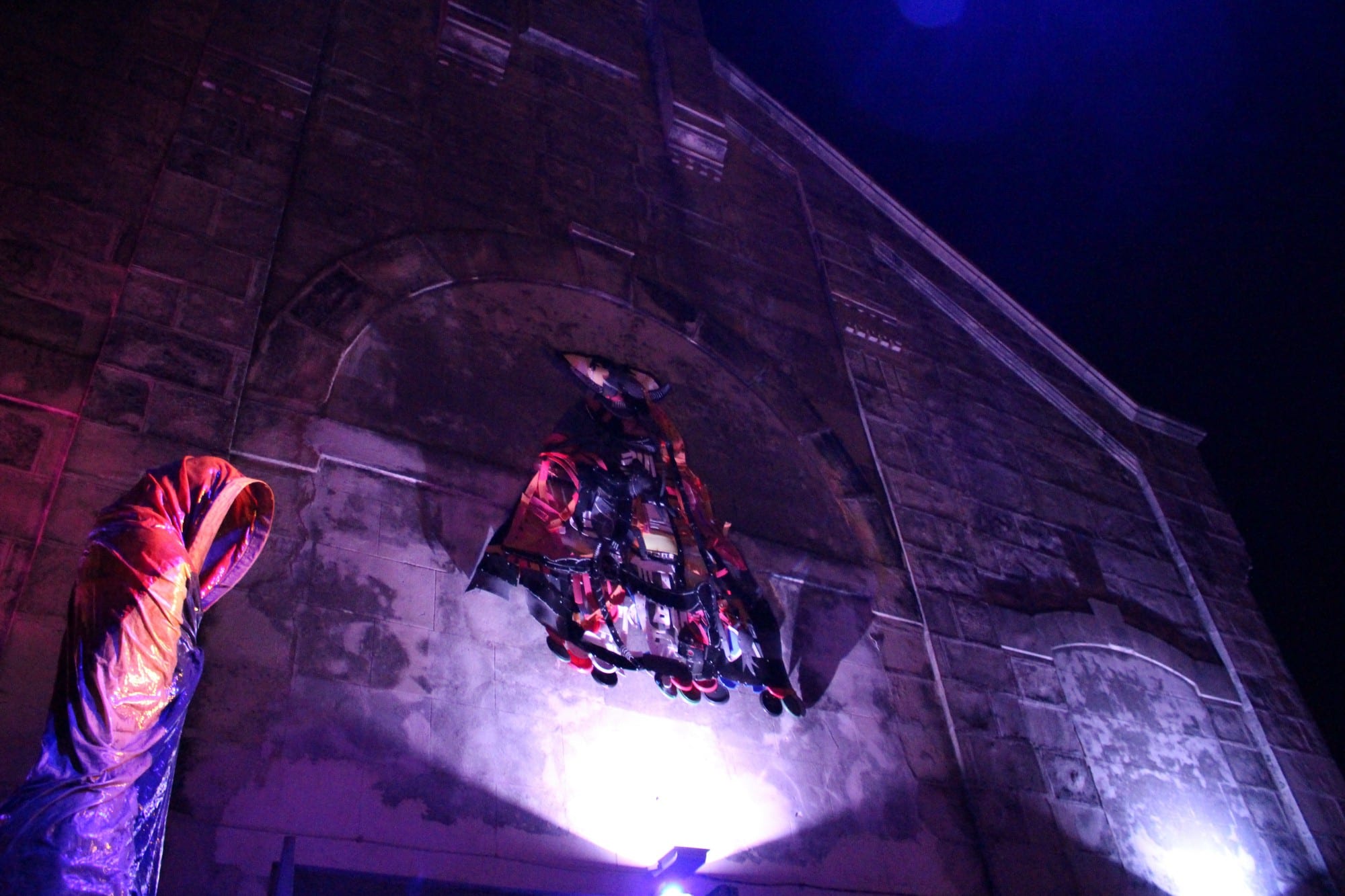 What an interesting and unusual scene we're witnessing here. Is that a hooded figure to the left, and another figure possibly descending from above? Cuba Unusual Museum Image: The side of the building is lit from below. There appears to be a hooded figure to the left, and another figure possibly descending from above.