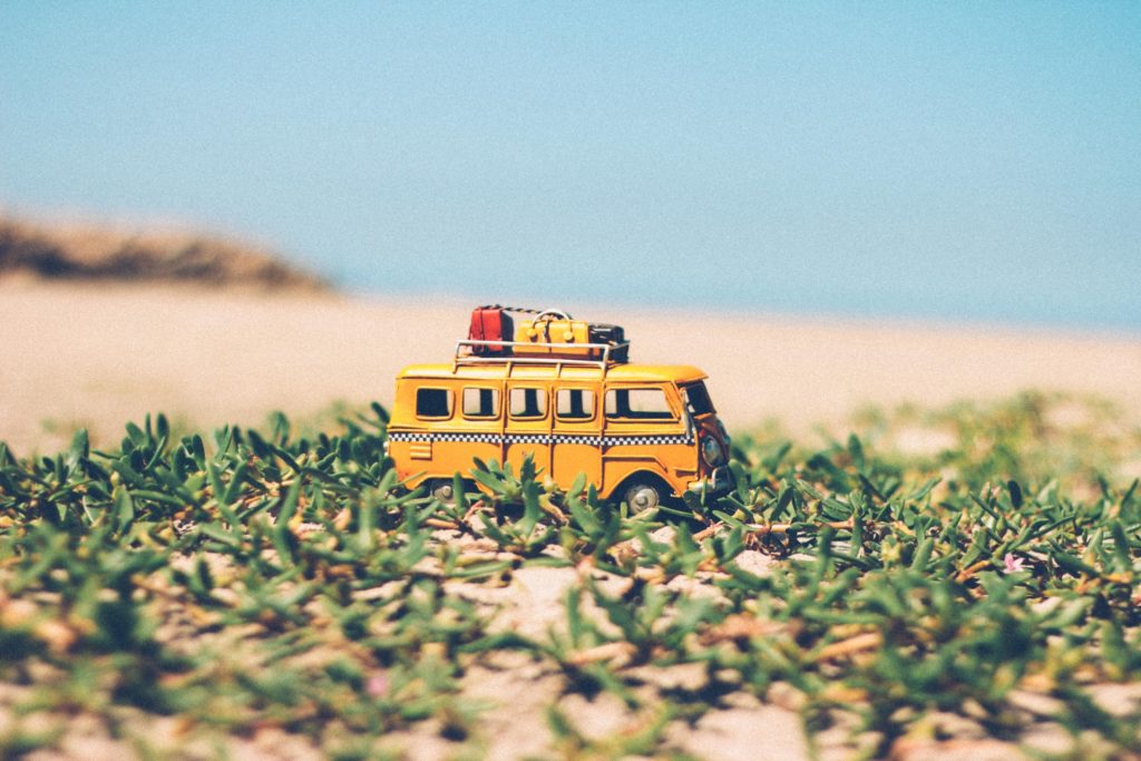 Beep-beep! Let's get going! Family Adventure Image: A toy VW van painted to look like a classic yellow taxi (with black and white accents) sits on a grassy patch of a sandy beach. Atop the toy van is a luggage rack containing toy suitcases in red, yellow, and black.