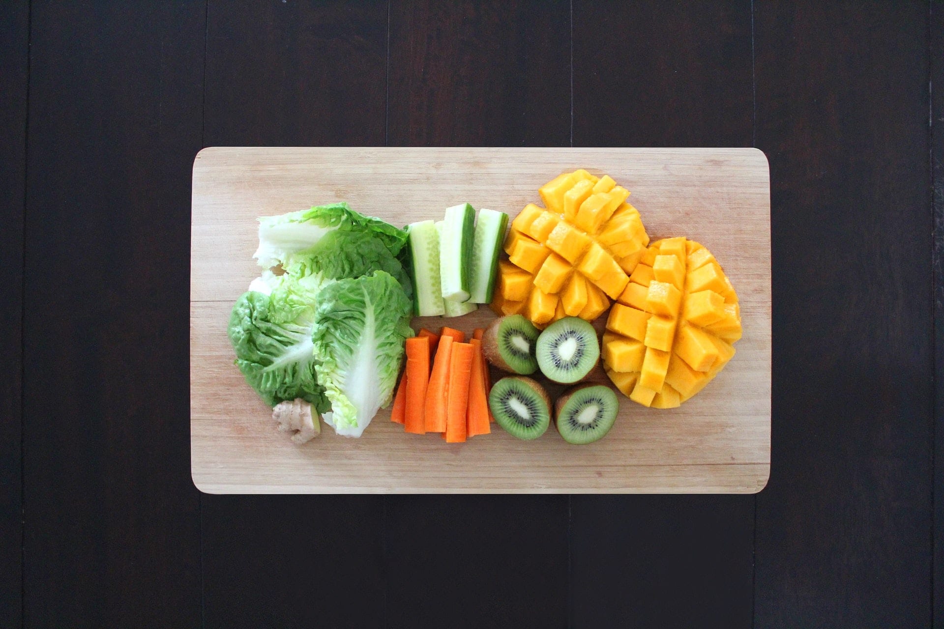 Family Adventure Image: There is a dark wood background. On a light wooden cutting board sits leaves of romaine lettuce, a piece of ginger, carrot sticks, cucumber sticks, kiwi halves, and mangoes turned inside out and cubed. 