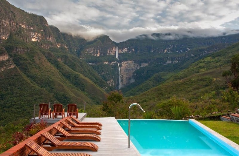 Best Hikes In Peru Image: A hotel swimming pool is surrounded by wooden lounge chairs, and offers an incredible view of the waterfall at the other end of this mountainous landscape.