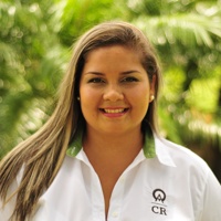 Katia Quirós is pictured wearing small earrings. Her dark blonde hair is draped over her shoulder, and she has a bright smile on her face.