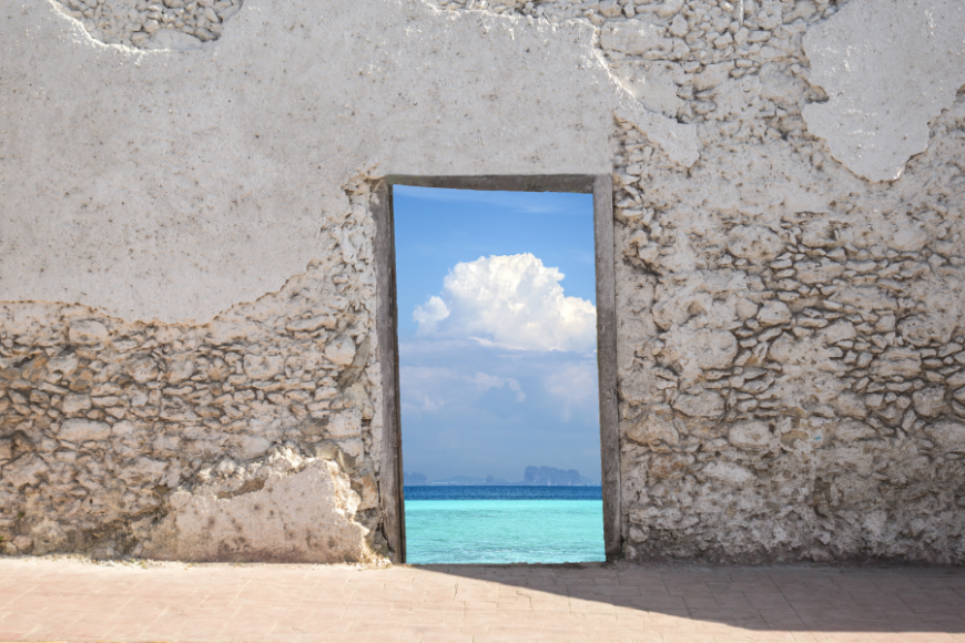 Moving Abroad Image: A weathered wall has a doorway cut into it, leading to a blue sea and sky.