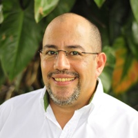 The distinguished Pablo smiles whilst wearing glasses and a white button-down shirt.