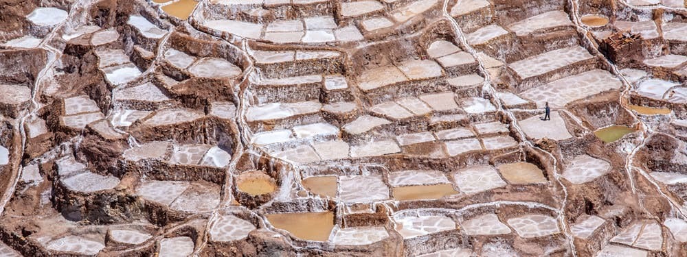Best Hikes In Peru Image: A photograph taken from an elevate view shows a person exploring the salt mines, some of which are still filled with water.