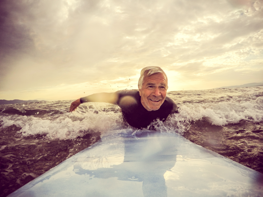 Retiring Abroad, Moving Abroad Image: A silver haired retiree wearing a wetsuit enjoys his surf board.