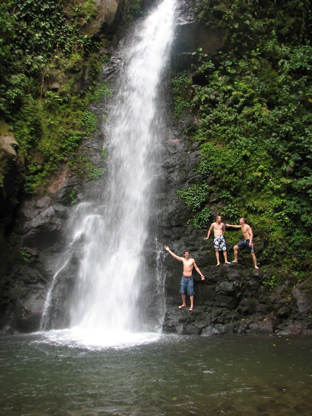 Swimming Holes Image: Three young men in swim shorts pose near the foot a swimming hole that has a waterfall pouring into it.