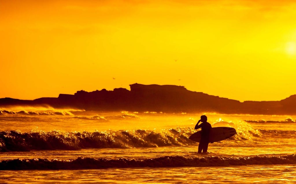 Destinations Worth Dreaming Image: A surfer stands in the ocean observing waves. We see little more than the silhouette because the sunlight is faded, and bathing everything in shades of orange and gold.