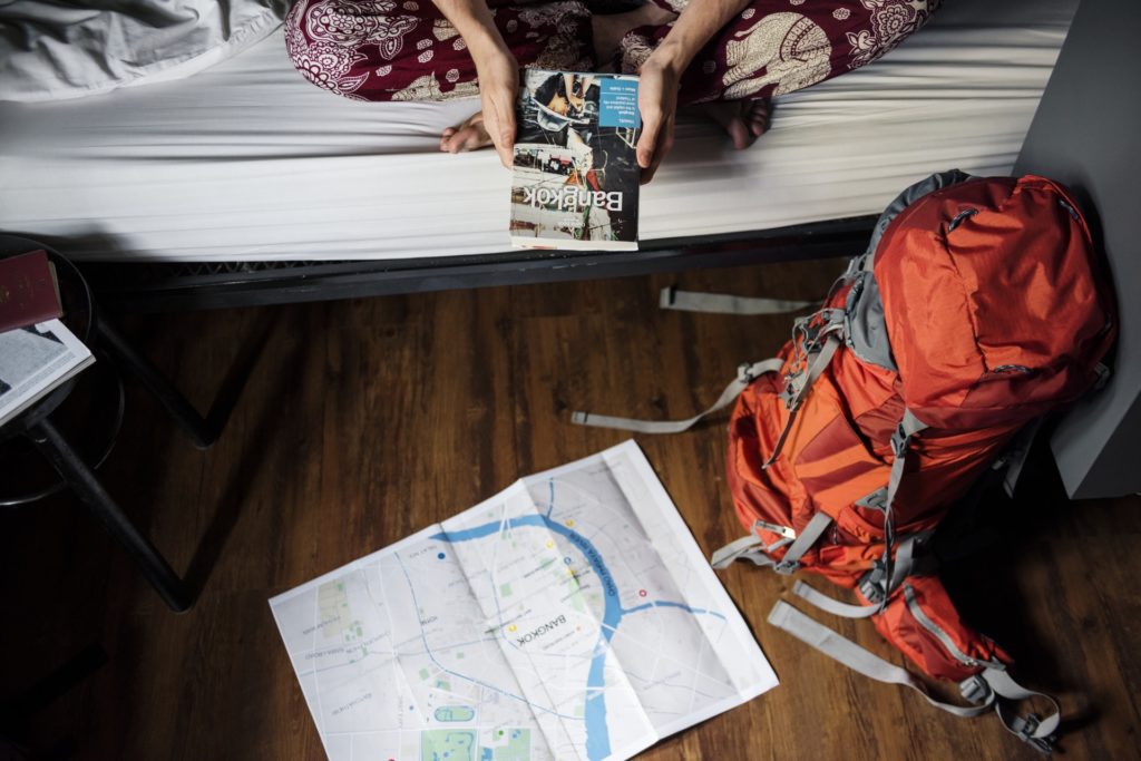Legally Travel To Cuba Image: A student wearing printed pants sits on a bed holding a travel book. An orange backpack sits in the right-hand corner of a wooden floor; a map is open on the floor as well. On the left-hand side of the room is a black stool with papers on top. 