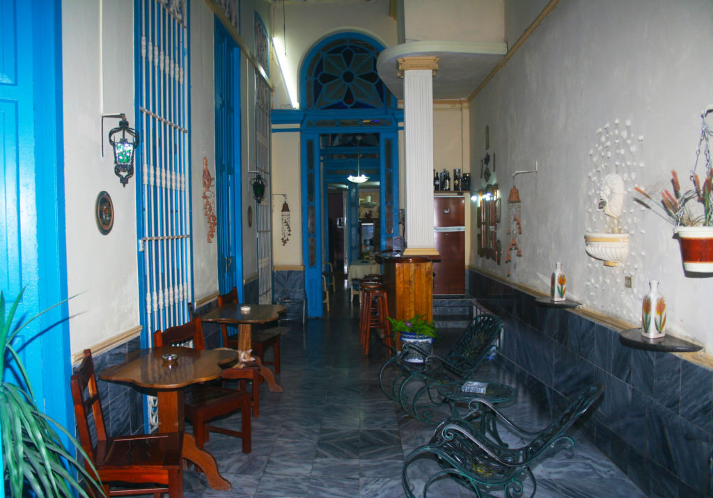 Legally Travel To Cuba Image: Another interior view of Casa Pardo Colonial—this time showing a room (which is almost like a very wide hallway) with bright blue door and window accents filled with tables and chairs.