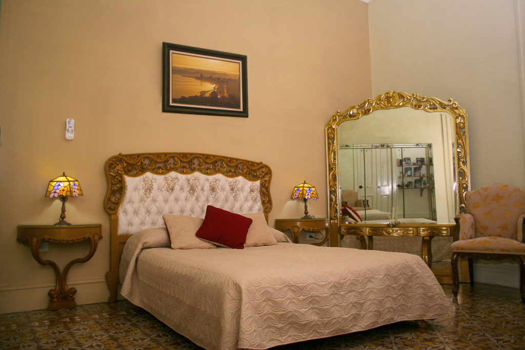 Legally Travel To Cuba Image: A simpe but well decorated room in the Hostal del Angel includes a large bed, two nightstands with lamps. an oversized mirror, and chair.