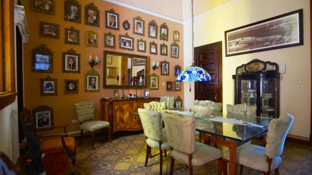 Legally Travel To Cuba: Another room in the casa particular Hostal del Angel; a glass dining table has six chairs, and a wall of photos, four other chairs, and additional furniture.