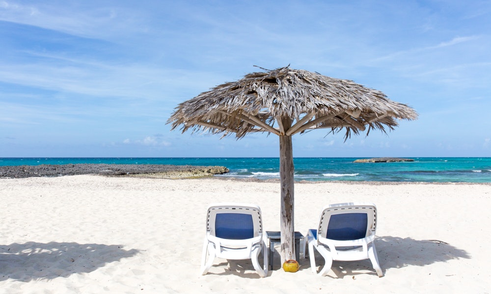 Legally Travel To Cuba: On a white-sand beach with a small crest of vibrant blue ocean water in the background sit two chairs, a table, coconut drink with straw, and a thatched beach umbrella. 