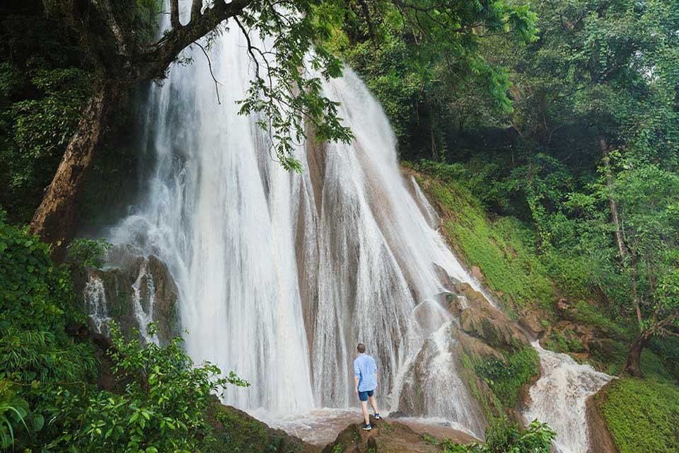 Destinations Worth Dreaming Image: A male traveller wearing dark-blue shorts and a light-blue shirt stands on a rock observing a waterfall as it rushes and breaks down a slope in, streaming into white channels.